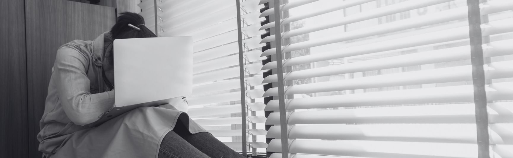 Woman hiding her face behind a laptop by a window, looking stressed